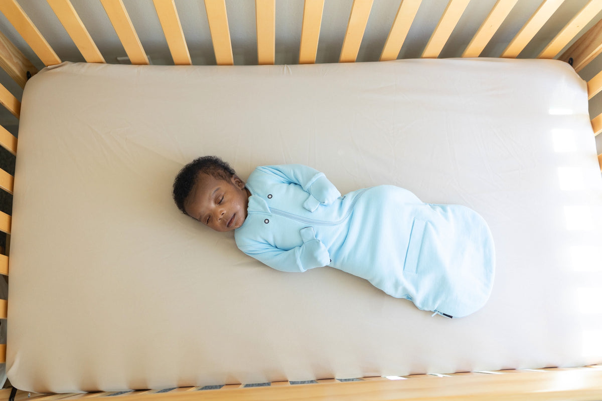Newborn asleep on their back in an empty crib, in a Sky Blue Swaddle Easy with the mittens closed