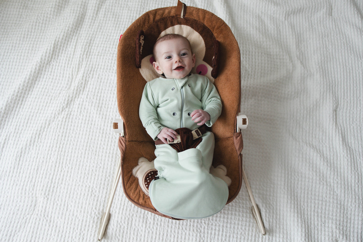 Smiling baby in a Sage Green Swaddle Easy in a bouncer, straps through the pocket between the legs