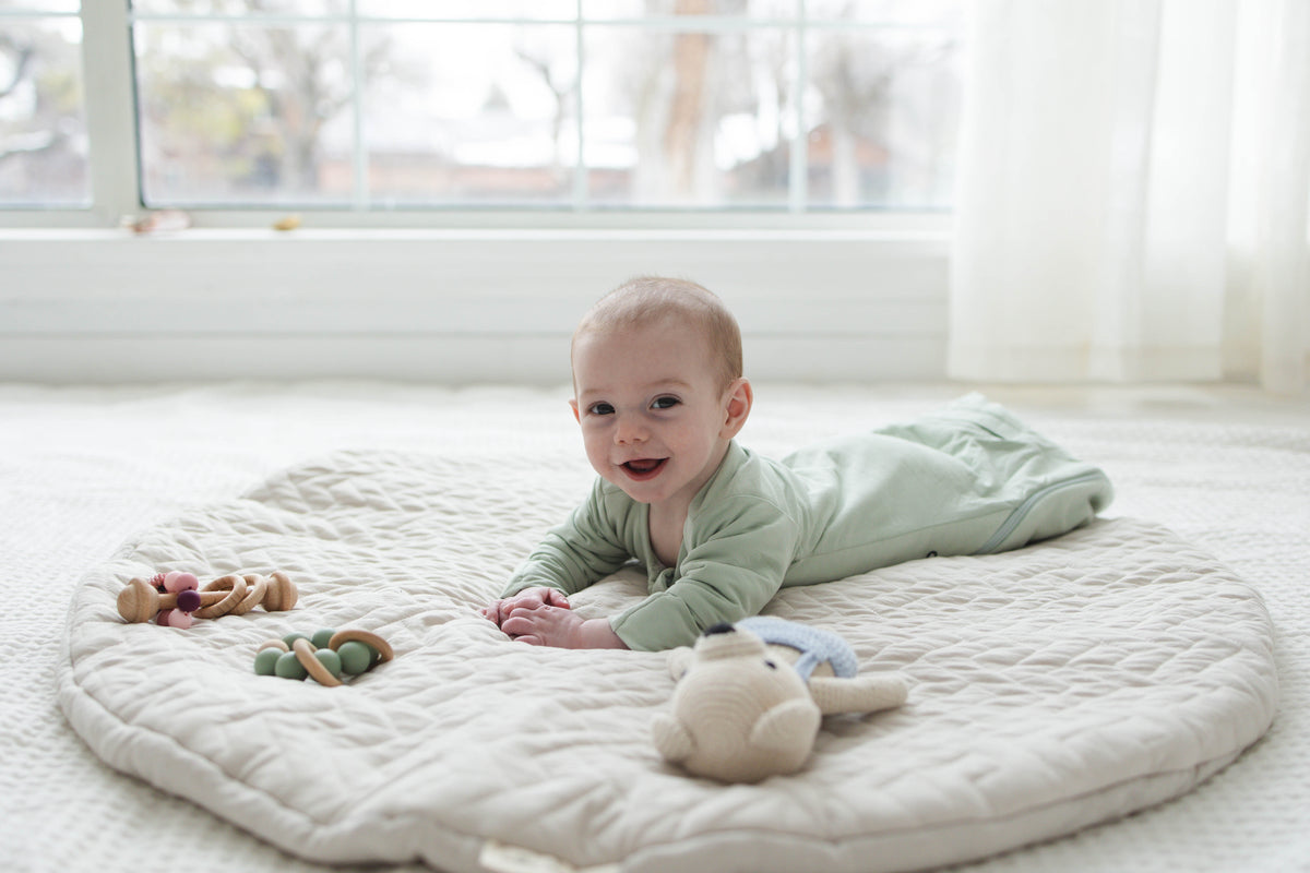 Smiling awake baby doing tummy time on a play mat in a Sage Green Swaddle Easy, hands out