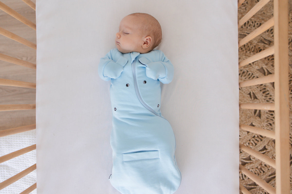 Baby asleep on their back in a Sky Blue Swaddle Easy in an empty crib, mittens folded over