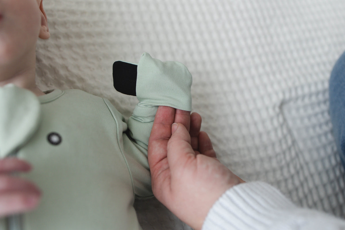 A parent folds a Sage Green mitten over a baby’s hand, the magnet tab facing out