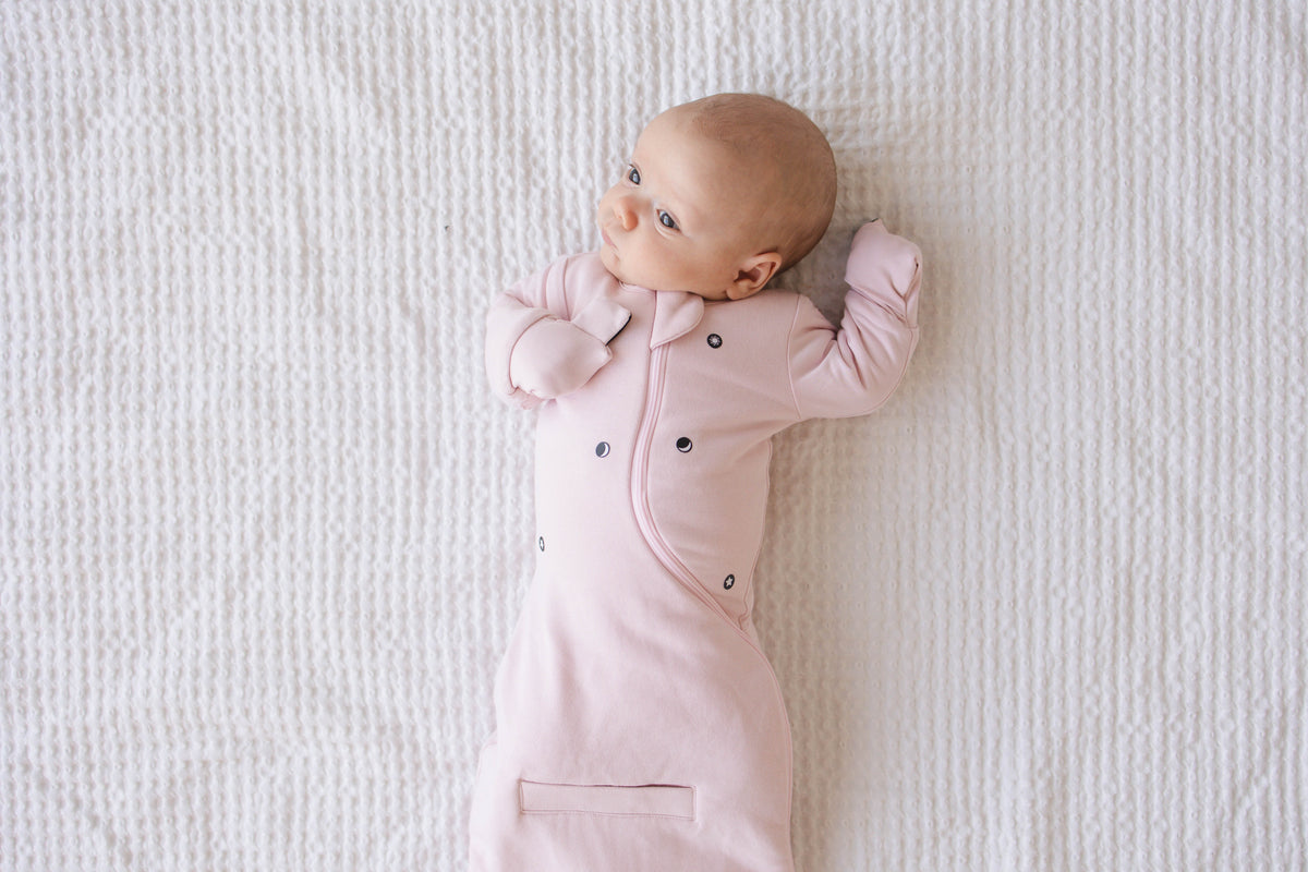 Awake baby in a Light Pink Swaddle Easy on a white blanket, mittens folded over