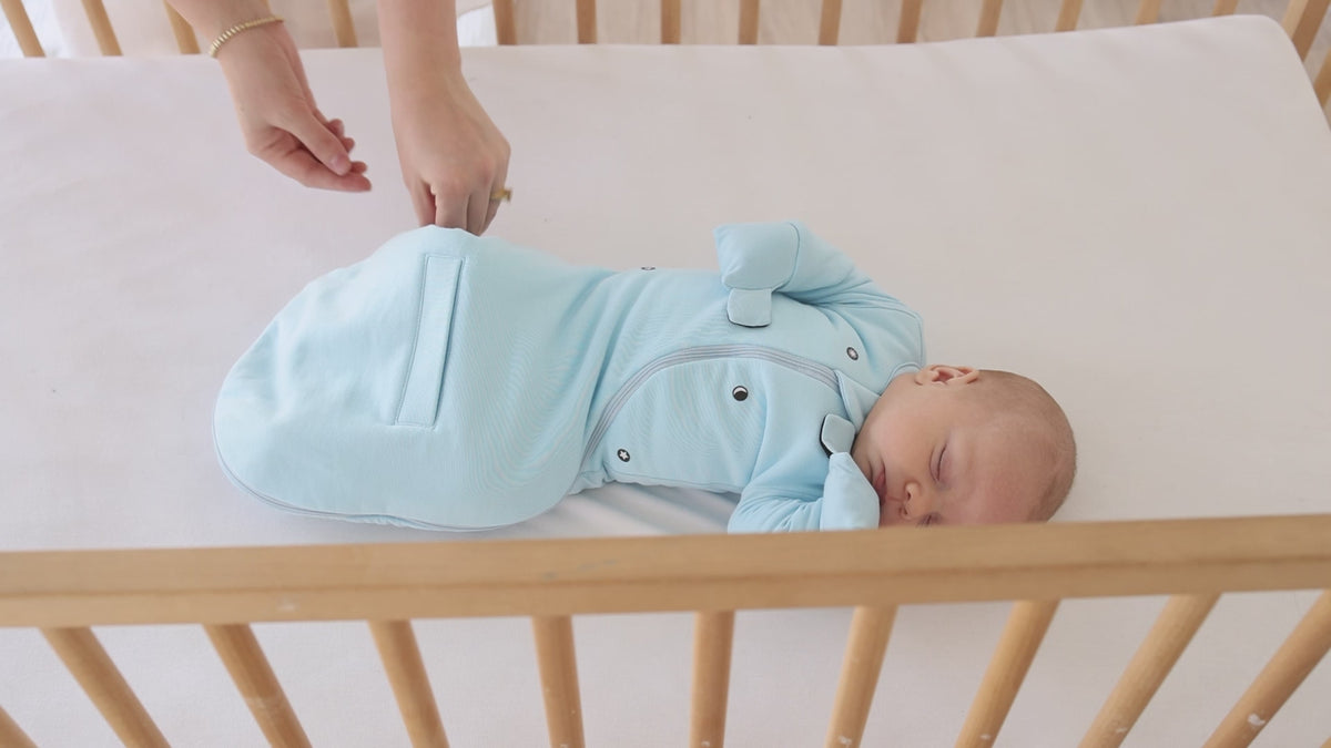 A parent opens the bottom zipper of a Sky Blue Swaddle Easy for a diaper change, baby in a crib
