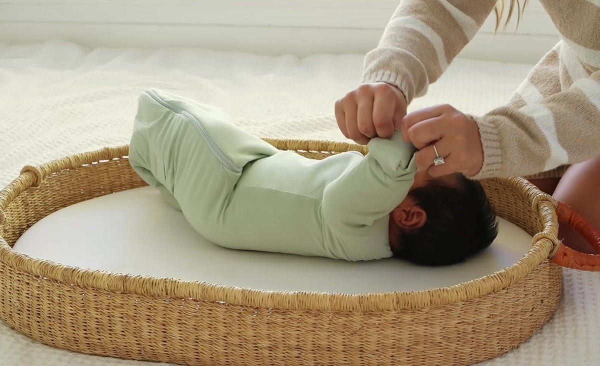 A mom folds the mittens over on a sleeping baby in a Sage Green Swaddle Easy, in a basket