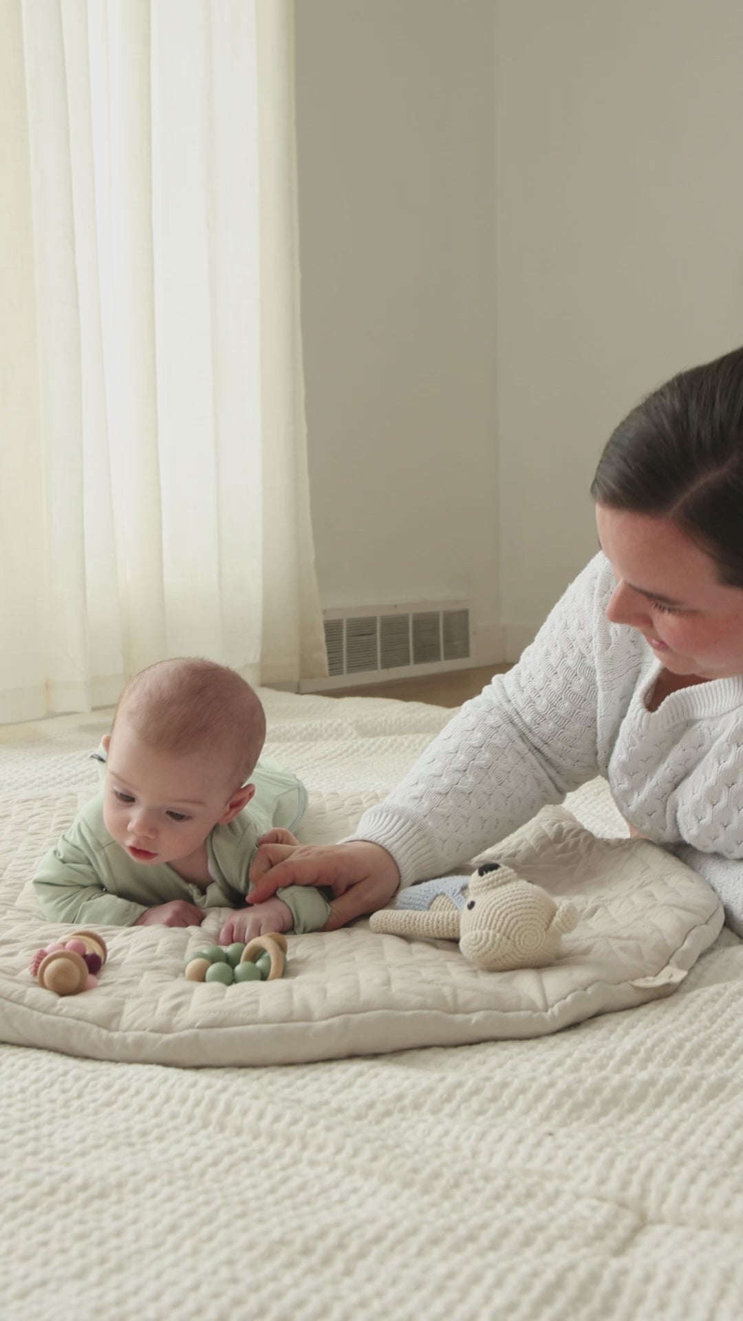 Awake baby on a play mat in a Sage Green Swaddle Easy with hands out, mom beside them