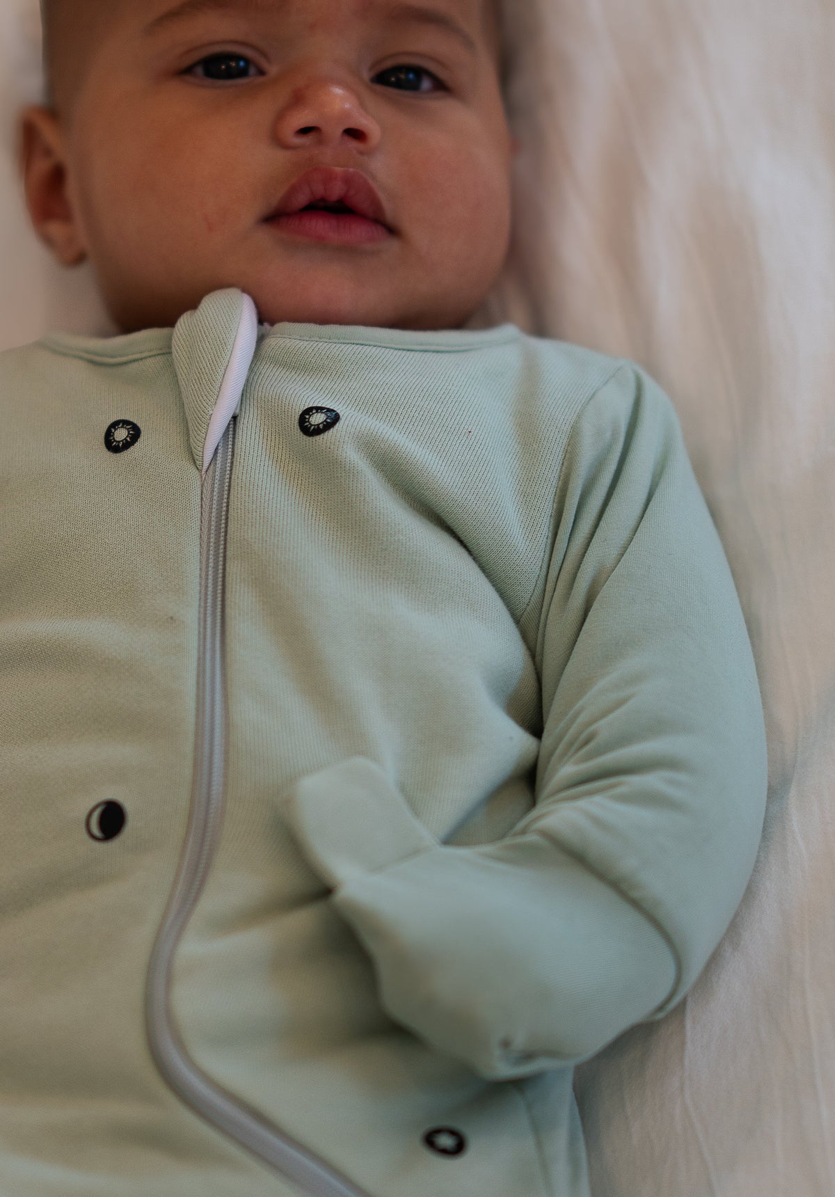 Awake baby in a Sage Green Swaddle Easy, close-up of a folded mitten, the magnet dots and the zipper