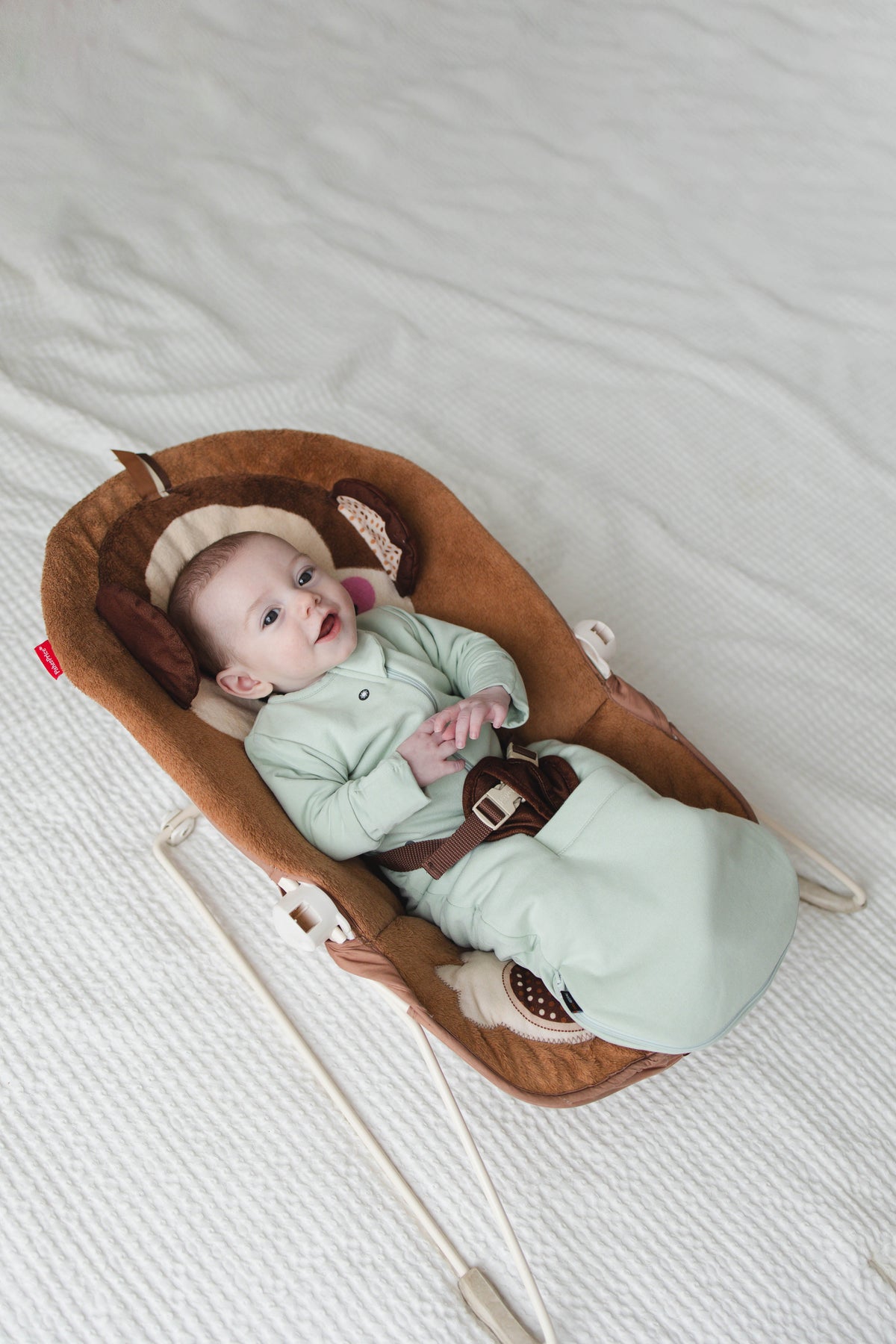 Smiling awake baby in a Sage Green Swaddle Easy in a bouncer, the straps through the pocket between the legs