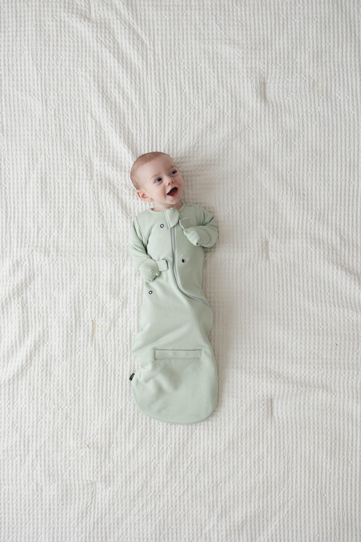 Smiling baby on their back in a Sage Green Swaddle Easy, snug with the mittens folded over