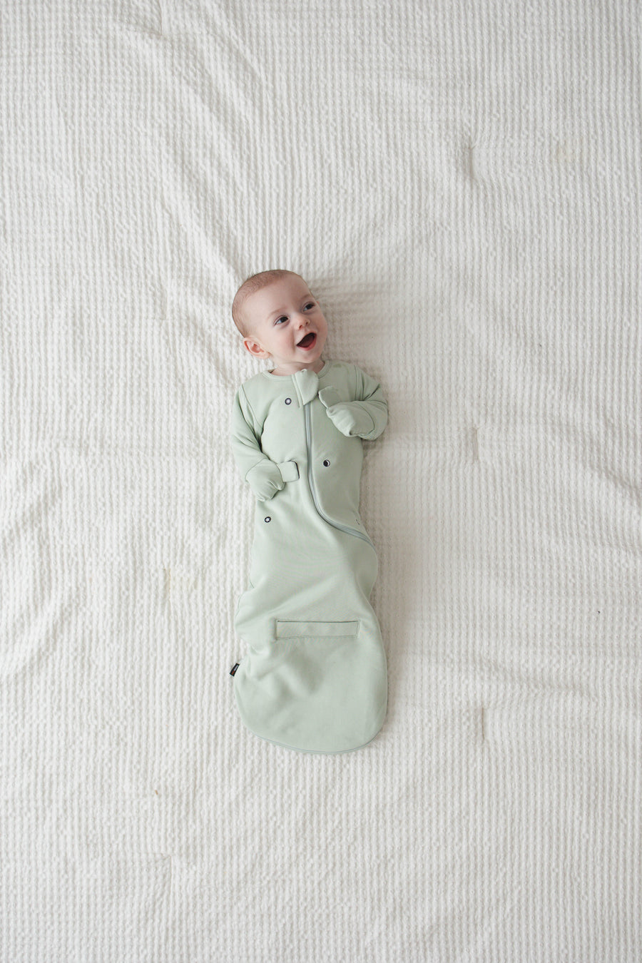 The same smiling baby on their back, snug in a Sage Green Swaddle Easy with the mittens folded over
