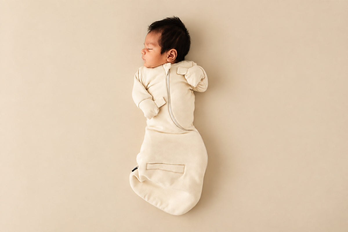 Newborn asleep on their back in a Cream Swaddle Easy, mittens folded over with hands up, on a beige backdrop