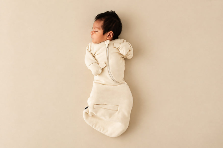 Newborn asleep on their back in a Cream Swaddle Easy, mittens folded over with hands up