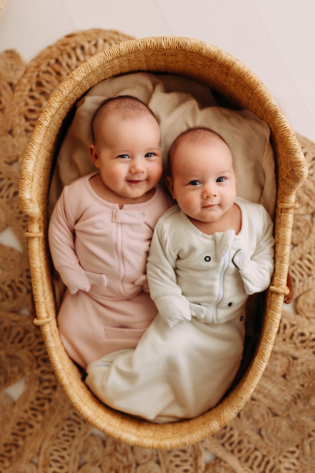 Smiling twin babies side by side in a woven basket, one in a Light Pink and one in a Cream Swaddle Easy, mittens folded over